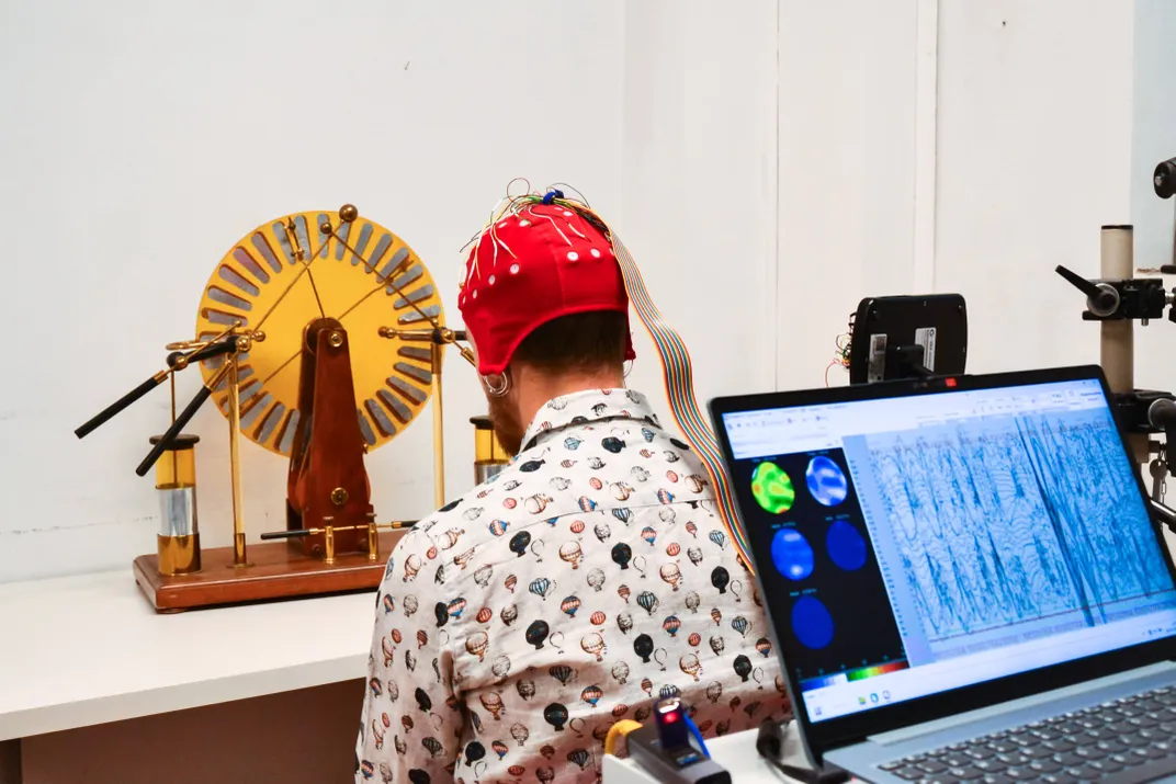 Eine Person sitzt mit dem Rücken zur Kamera und blickt auf ein wissenschaftliches Instrument wie ein erhöhtes Rad, eine Kappe mit Elektroden sitzt auf ihrem Kopf und ein Computerbildschirm zeigt die Gehirnaktivität