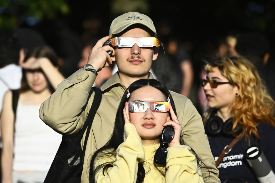 Zwei Personen blicken mit einer Sonnenfinsternisbrille in die Kamera, die Sonne wird reflektiert