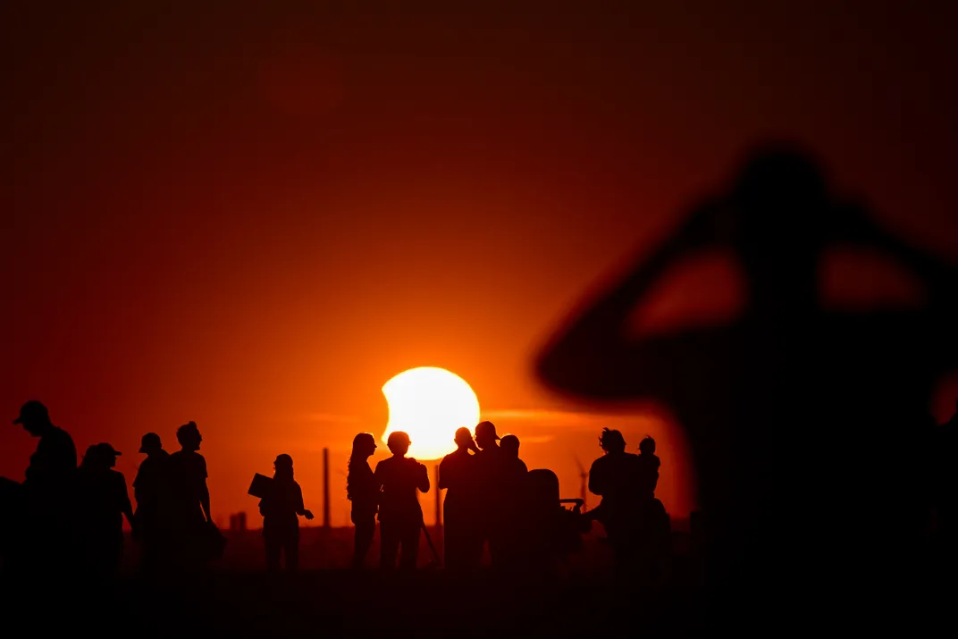 links eine tief hängende Sonne, von der ein kleiner Teil vom Mond verdeckt ist, dahinter eine Menschenmenge als Silhouette