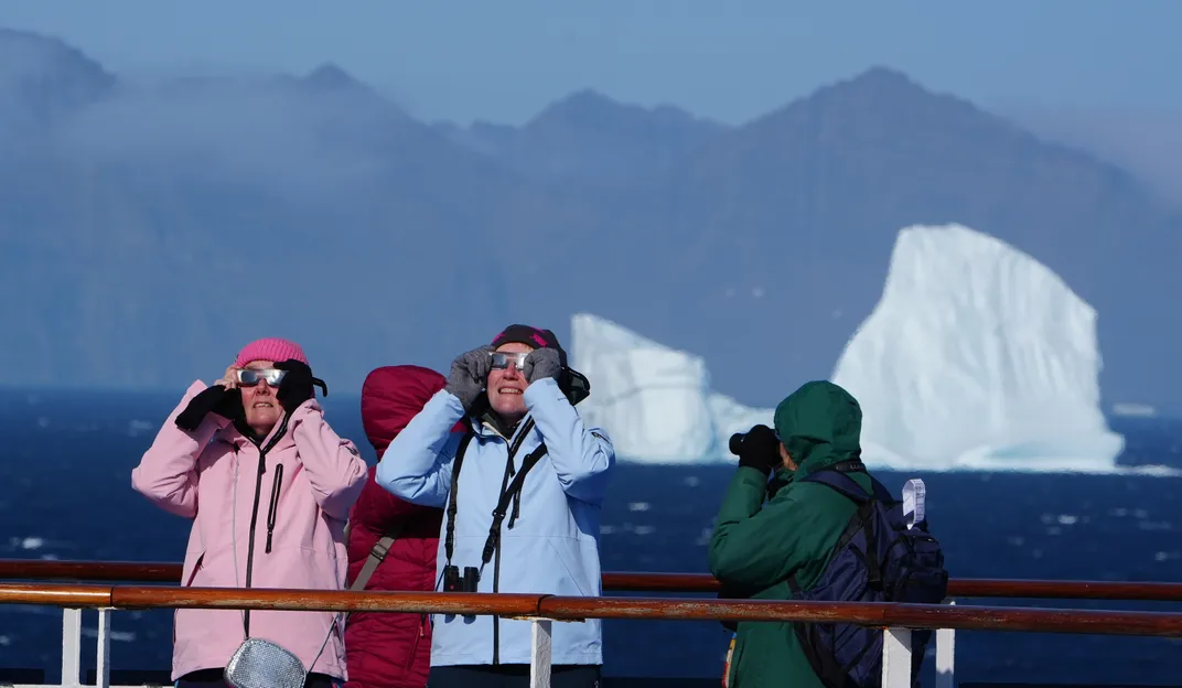 Menschen, die große Jacken tragen, benutzen eine Sonnenfinsternis-Brille mit einem Eisberg im Hintergrund