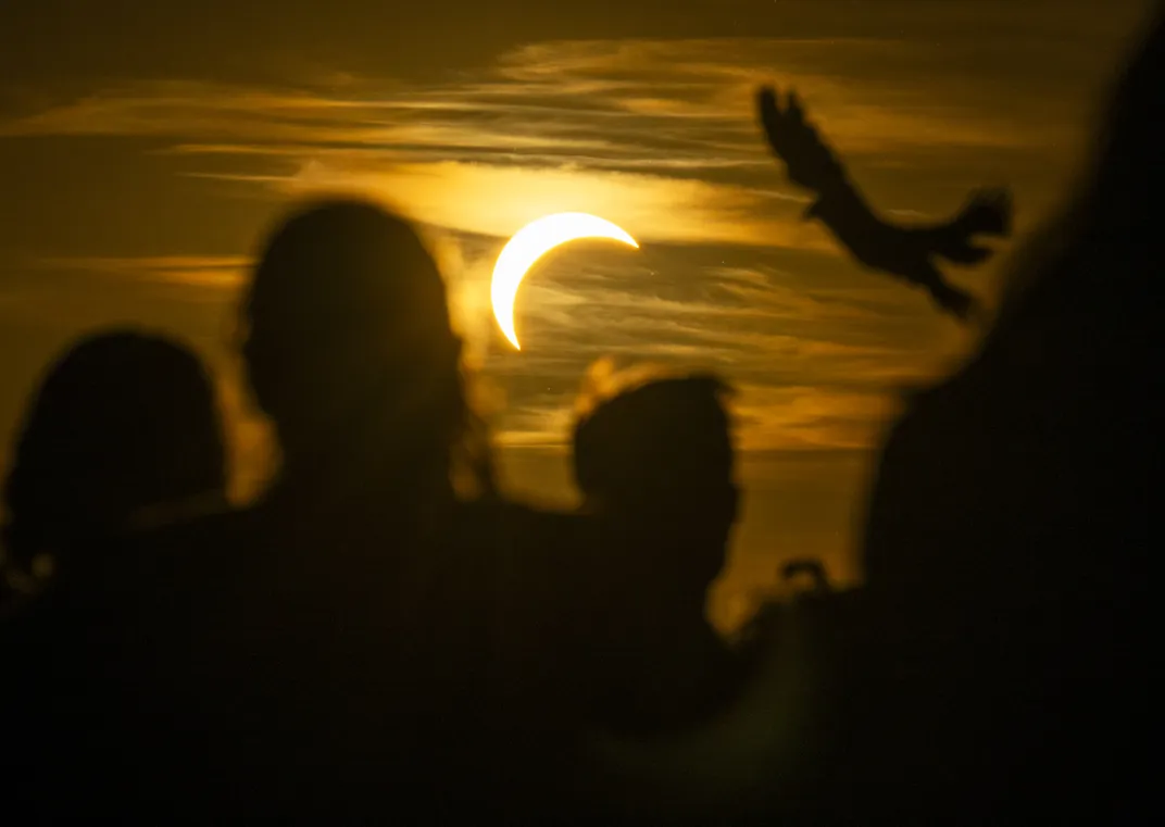 Eine teilweise verfinsterte Sonne steht hinter Wolken, während im Vordergrund Silhouetten von Menschen und einem fliegenden Vogel zu sehen sind