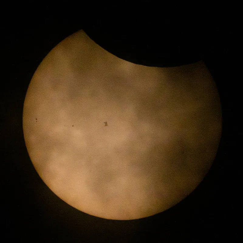 Die Oberfläche der Sonne sieht aus wie eine schwach orangefarbene Kugel, an deren Spitze der Mond etwa zehn Prozent verdeckt, und die Silhouette eines Raumfahrzeugs bedeckt einen Teil seiner Oberfläche